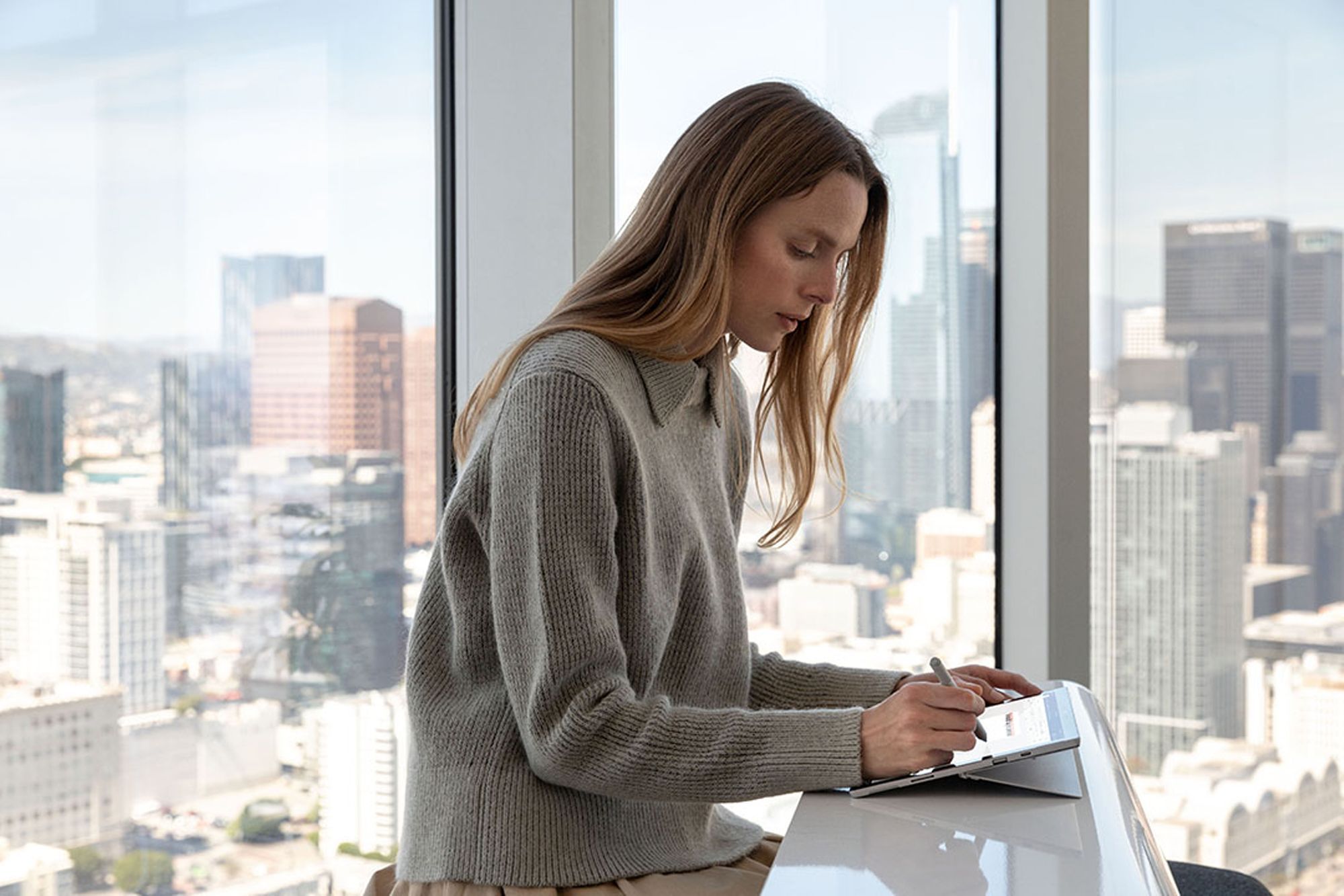 En kvinde sidder ved et bord foran en vinduesvæg, bag hvilken en storby kan ses. Hun arbejder med Surface Pen på Surface Pro 7