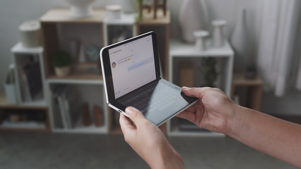 A person is holding the Surface Duo. One screen is used as a keyboard, the other one shows a chat.