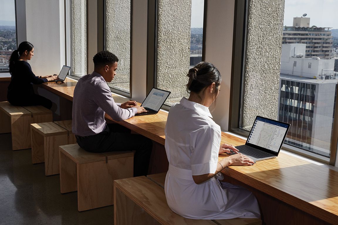 In einem Büroraum mit großen Fenstern sitzen drei Personen an Schreibtischen und arbeiten auf ihren Surface Laptops 