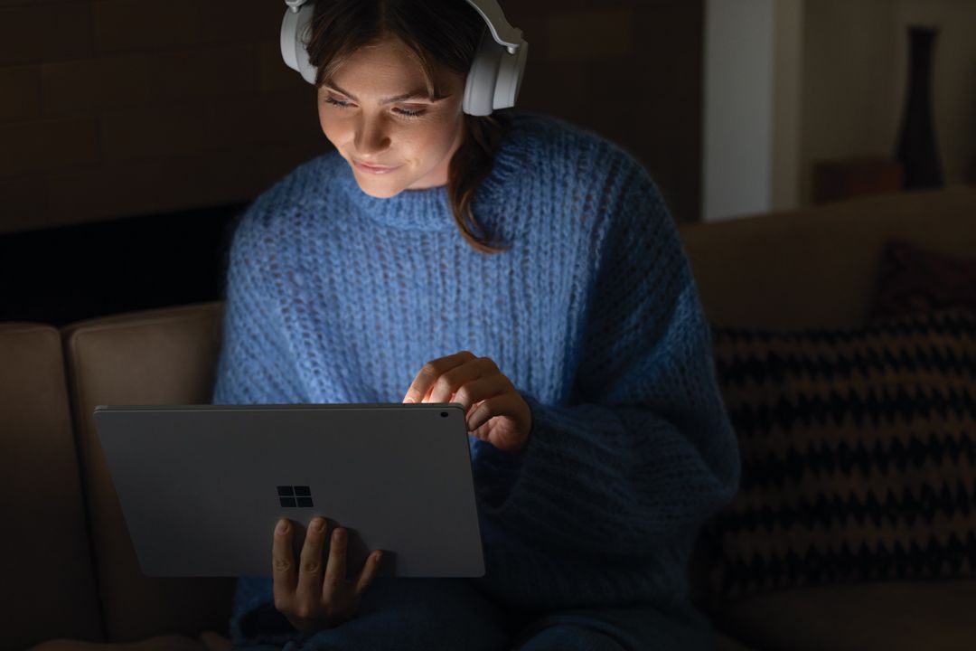 Eine Frau sitzt auf einer Couch, trägt Surface Headphones auf dem Kopf und schaut sich etwas auf dem Surface Book an