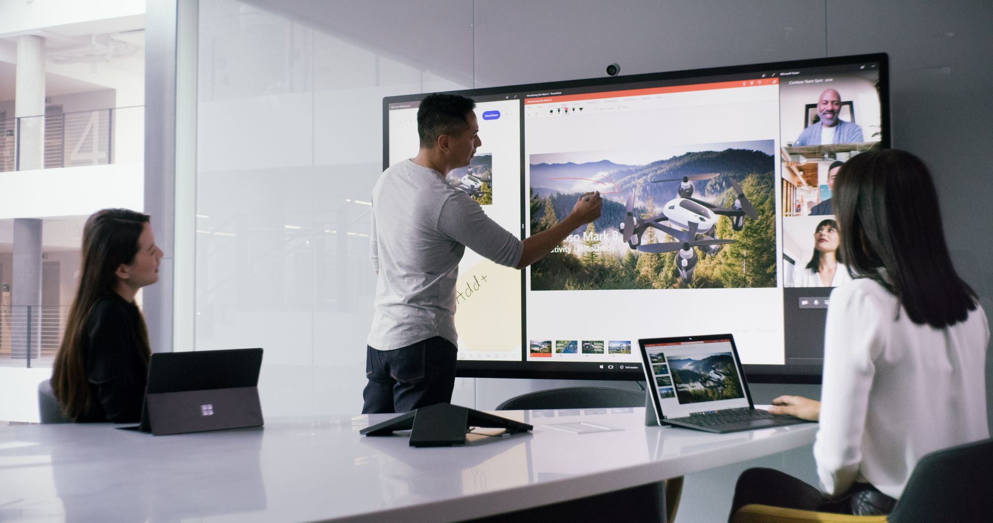 In a meeting room, two women are sitting at a conference table with their Surface devices and a man is standing at the Surface Hub, working on a presentation with the Surface Hub 2 Pen