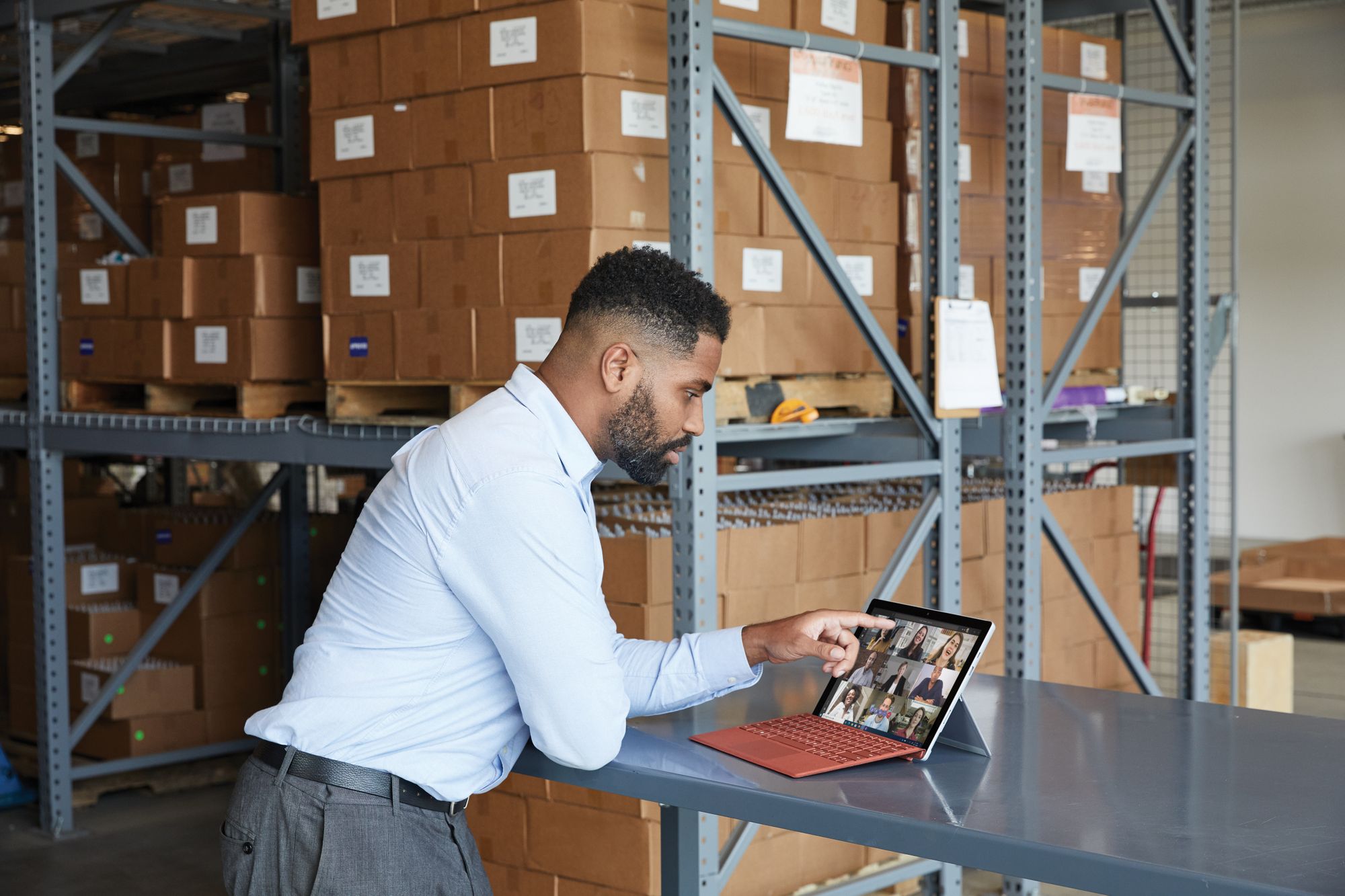 In a warehouse, a man stands at a table looking at the Surface Pro 7 Plus in laptop mode, which is showing a video conference