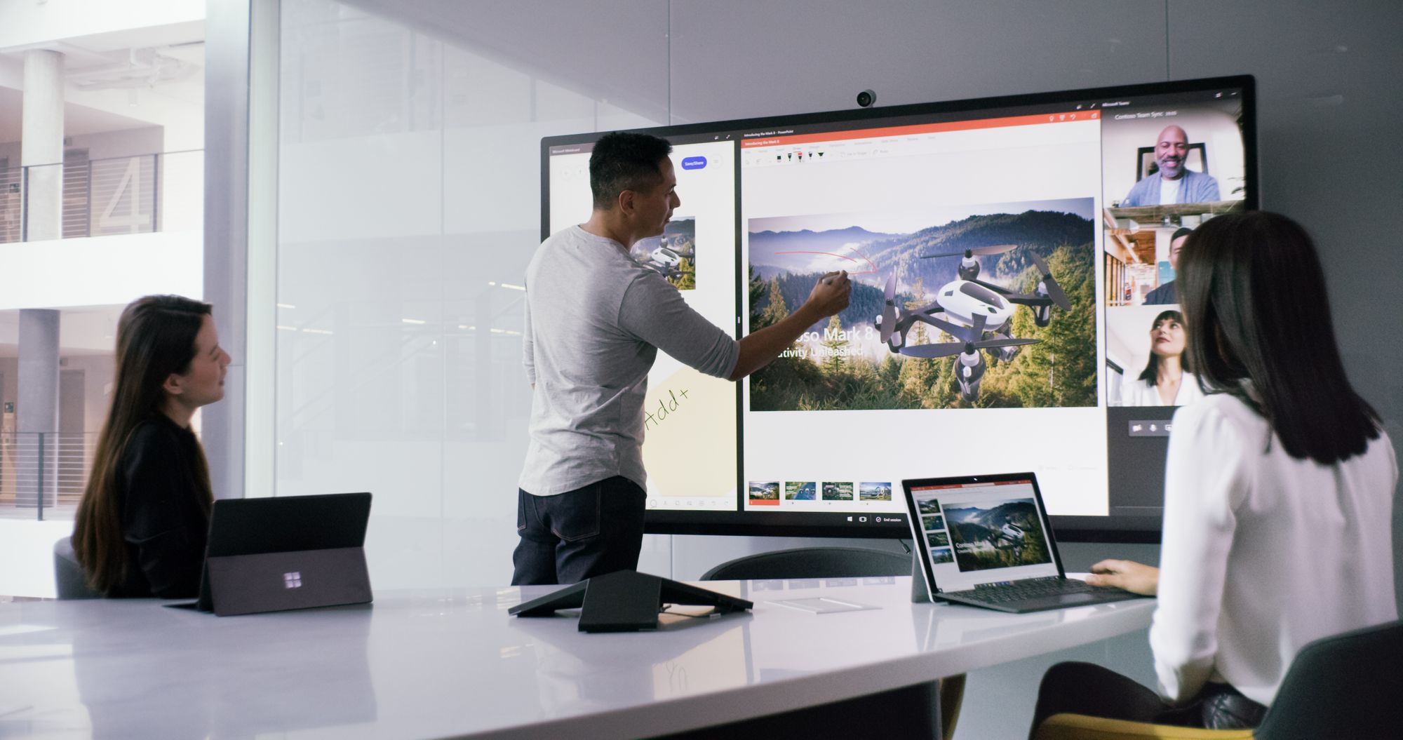 I et mødelokale sidder to kvinder ved et konferencebord med deres Surface-enheder, og en mand står ved Surface Hub, hvorpå han holder en præsentation med Surface Hub 2 Pen 