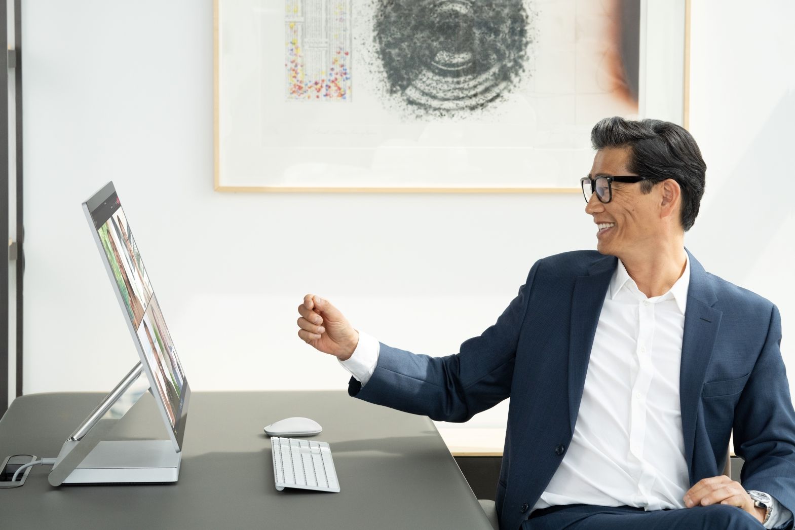 Man sitting at his desk, smiling with his hand outstretched towards his Surface Studio 2+ while on a Teams video call